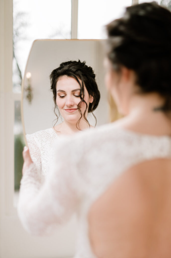 Bruid met speels opgestoken haar, natuurlijke make-up en een glanzende look, kijkend in de spiegel.